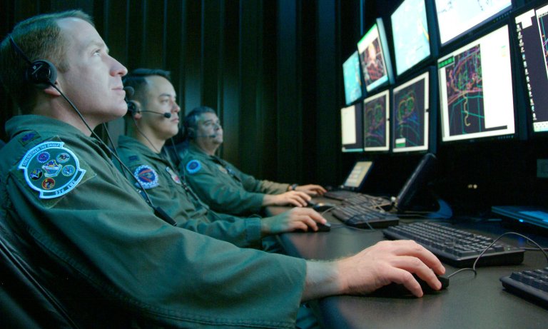 Three US air force servicemen sit in uniform in front of a bank of computers