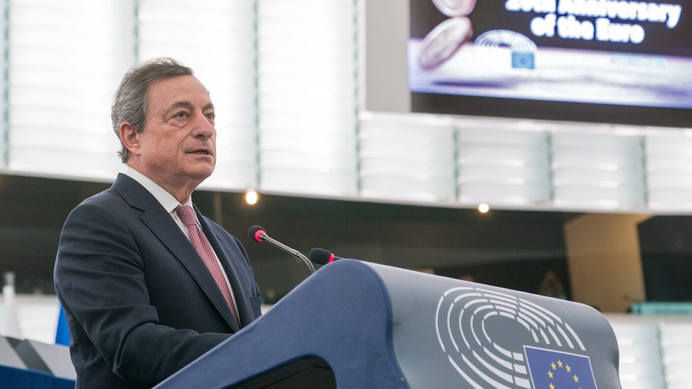 Mario Draghi speaks at a lecturn at the European Parliament, with a slide “20th Anniversary of the Euro"