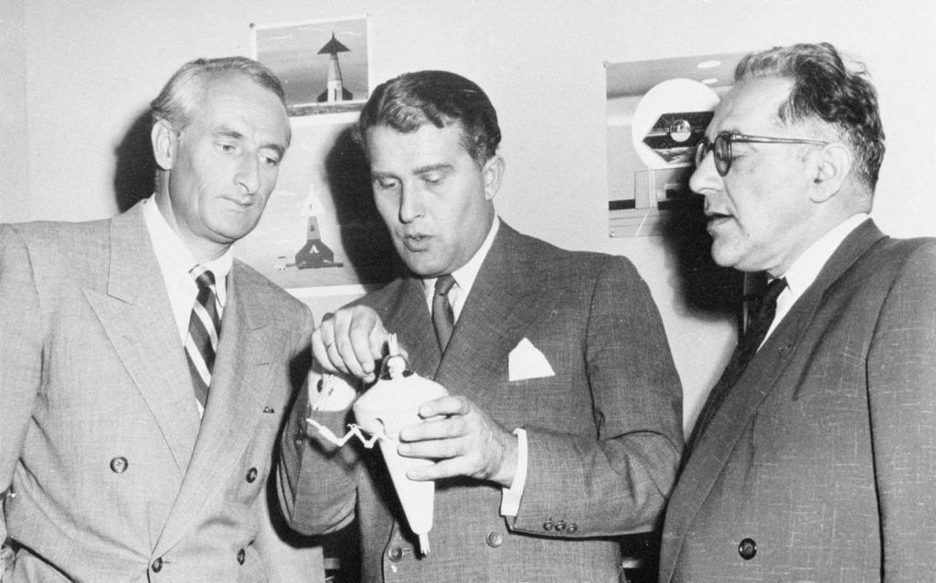 Three middle-aged white men (including the infamous former Nazi rocket scientist, Werner von Braun) in grey suits look at a rocket model
